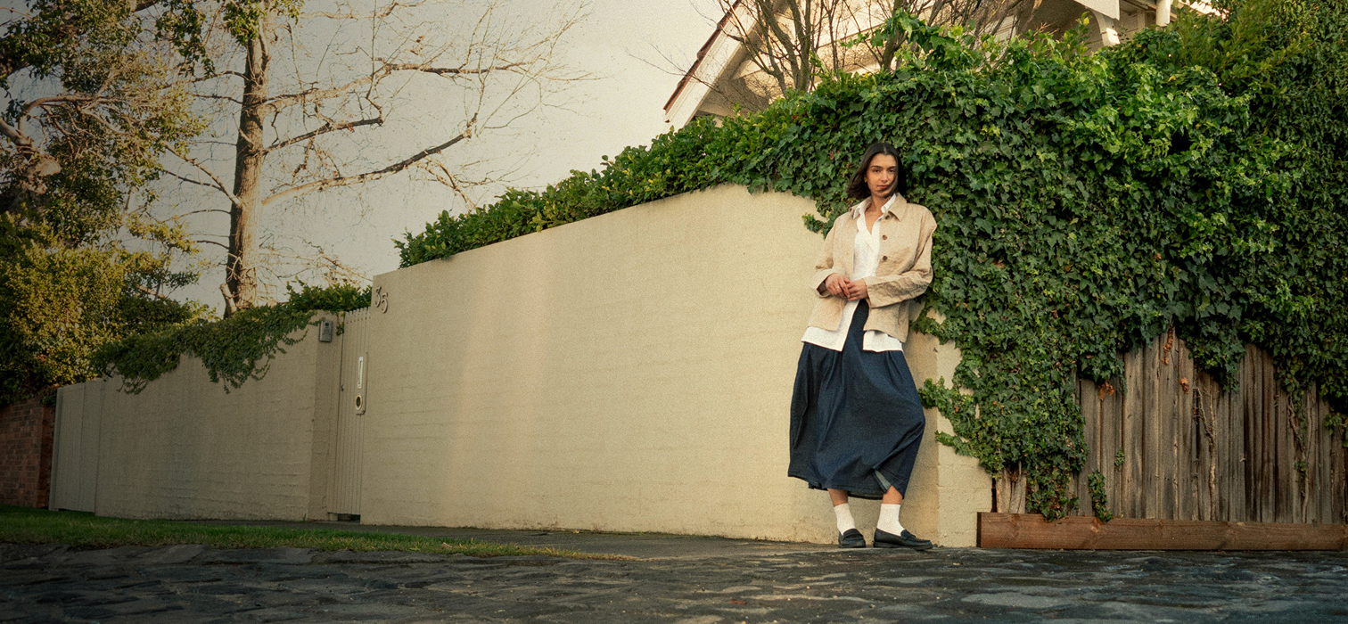 Woman standing against a wall with greenery, wearing a beige jacket and blue skirt.